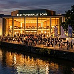 Beethovenhalle in Bonn am Abend: erleuchtete Glasfassade des Konzerthauses, Festivalpublikum auf dem Weg zum Konzert